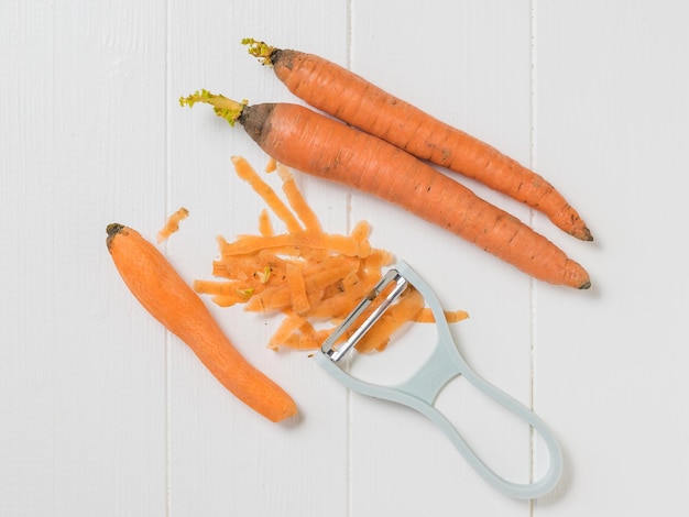 Tres zanahorias y un pelador sobre una mesa de madera blanca. Limpiar zanahorias con un cuchillo especial. Endecha plana.