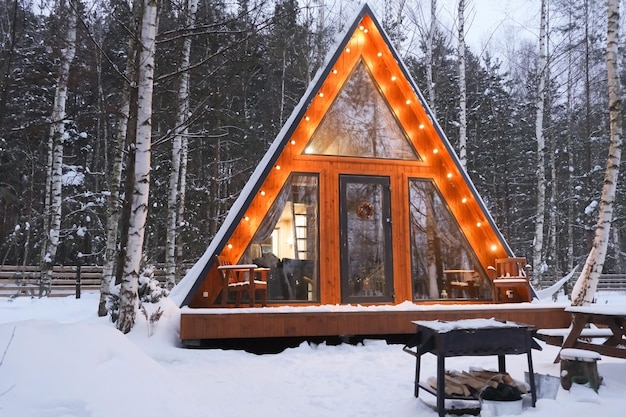 Triángulo moderno casa de madera en bosque de invierno entre árboles de campo con nieve blanca en el techo