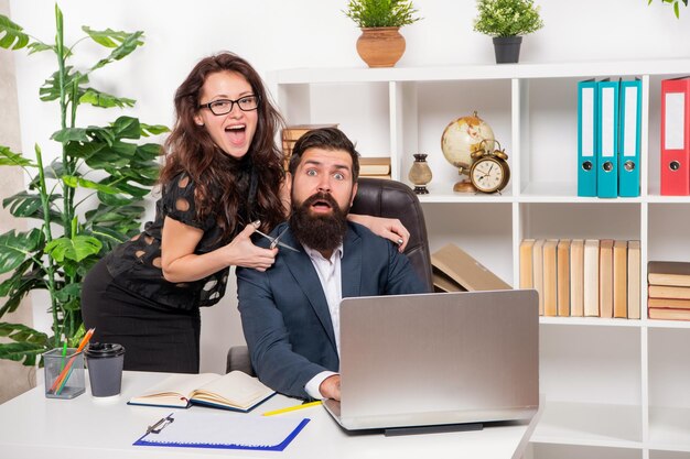 Tricky woman boss asustar a un hombre barbudo empleado cortando la barba con unas tijeras en la barbería de la oficina