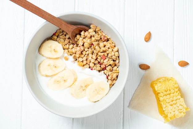 Trigo branco e granola com iogurte desnatado em uma tigela branca em uma composição com uma colher, favos de mel, banana em fundo branco de madeira.
