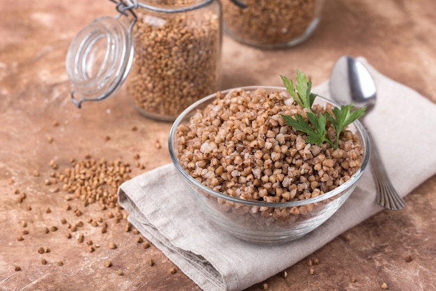 Trigo sarraceno cocido en un recipiente de vidrio, transparente, hoja de perejil, servilleta de lino, frascos para almacenar cereales, fondo marrón, contenido de salud, vista superior