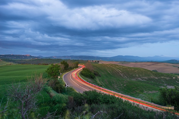 Foto trilha de luz de carro na estrada sinuosa na paisagem de colina única da toscana itália céu dramático do pôr do sol nas aldeias iluminadas ao crepúsculo no topo da colina