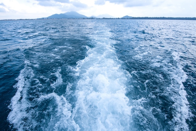Trilha e ondas espumosas na superfície do mar azul, atrás do barco em movimento com o fundo da ilha.