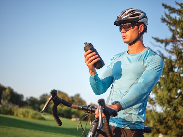 Trinken Sie durstigen professionellen männlichen Radfahrer, der eine Flasche mit Wasser hält, die mit seinem Fahrrad steht