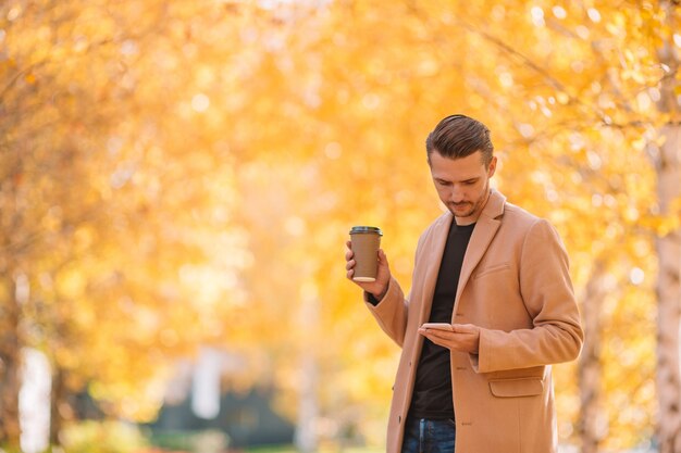 Trinkender Kaffee des jungen Mannes mit Telefon im Herbstpark draußen