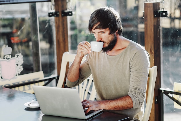 Trinkender Kaffee des jungen Mannes und Anwendung des Laptops Café im im Freien. Freiberufliche Konzeption