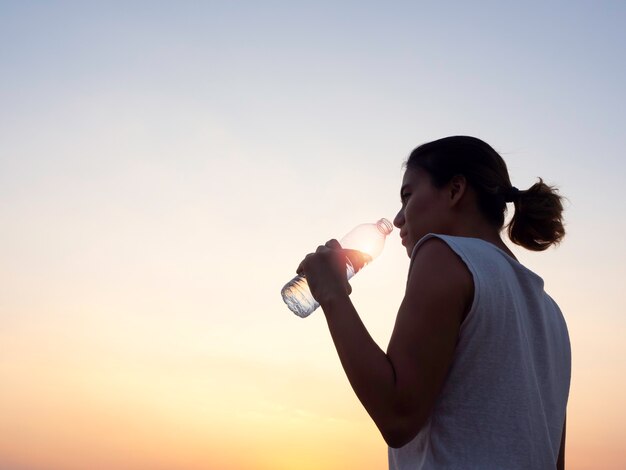 Trinkwasser der asiatischen Frau von der Plastikflasche nach dem Training, das am Abend-Sommer auf schönem Sonnenuntergangshimmel mit Kopienraum ausübt