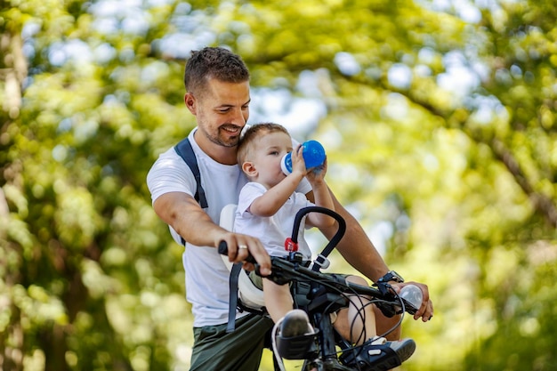 Trinkwasser und Radfahren Der Junge trinkt Wasser aus einer blauen Flasche, während Papa ihn ansieht