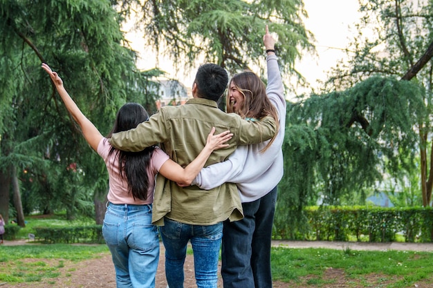 Trío alegre en el parque hombre asiático abrazado por mujeres uno apuntando hacia el cielo en ropa casual en medio de la vegetación