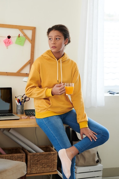 Triste adolescente de raza mixta en traje casual sentado en el escritorio con suministros y portátil y bebiendo jugo de taza en su habitación