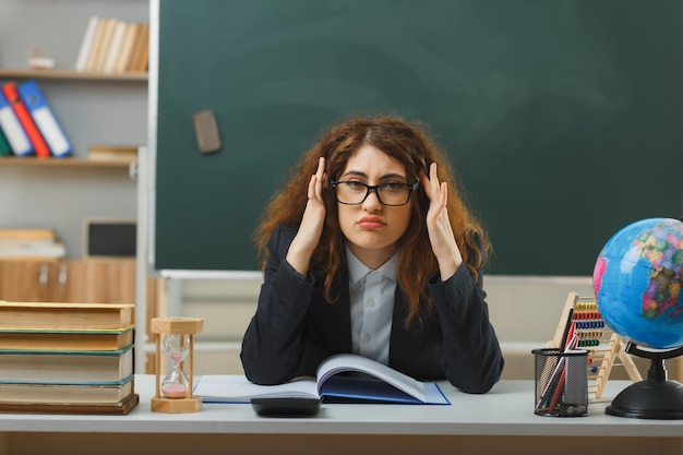 Triste agarró la cabeza de una joven maestra con gafas sentada en el escritorio con herramientas escolares en el aula