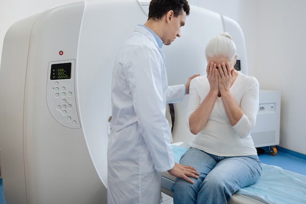 Triste anciana deprimida sentada en la mesa de examen del escáner CT y cubriéndose la cara mientras llora