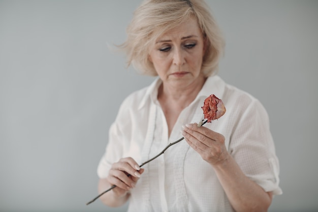 Triste anciana senior en camisa blanca sostiene marchita una vieja flor rosa muerta seca. Vejez y envejecimiento conceptual