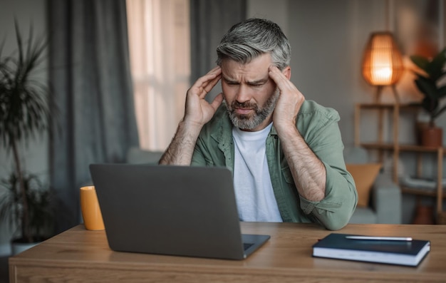 Triste anciano europeo con barba mira la computadora portátil en el lugar de trabajo sufre de dolor de cabeza en la sala de estar