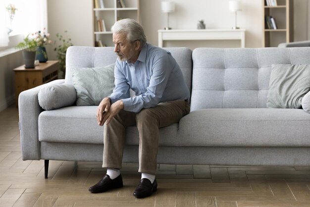 Triste anciano solitario de cabello gris sentado solo en el sofá de casa mirando hacia abajo en mal pensamiento sufriendo de depresión apatía lidiando con la pérdida malas noticias sobre problemas familiares de atención médica