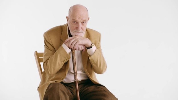 Triste anciano solitario sentado en un sillón con bastón aislado sobre fondo blanco.