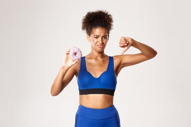 Triste atleta afroamericana en uniforme azul, mostrando sabroso donut y pulgar hacia abajo, estando en