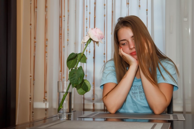 Triste bastante joven adolescente sentado a la mesa en casa con rosa solitaria, que simboliza la tristeza. Linda mujer muestra emociones actuando, en la ventana de la cocina en casa. Espacio de derechos de autor