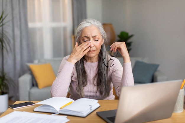 Triste cansada caucásica anciana canosa se quita las gafas y se limpia los ojos en el interior de la sala de estar