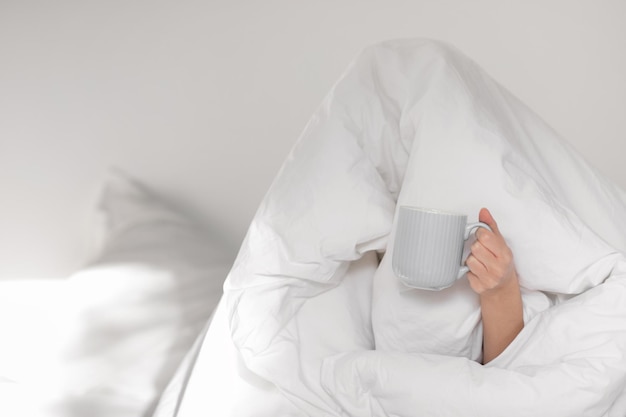 Triste, cansada y graciosa mujer caucásica milenaria sentada en la cama cubierta con una sábana blanca con una taza de café