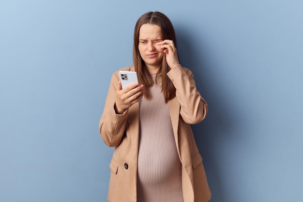 Triste y cansada joven adulta embarazada con vestido y chaqueta posando aislada sobre un fondo azul frotándose los ojos doloridos después de largas horas usando el teléfono inteligente haciendo caras de sentimientos desagradables