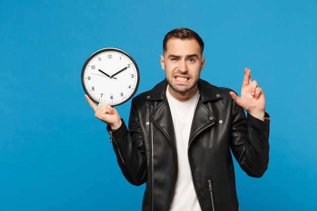 Triste chateado elegante jovem com barba por fazer na jaqueta de couro preta, camiseta branca, segurando o relógio redondo isolado no retrato de estúdio de fundo de parede azul. Conceito de estilo de vida de pessoas. Se apresse. Simule o espaço da cópia.