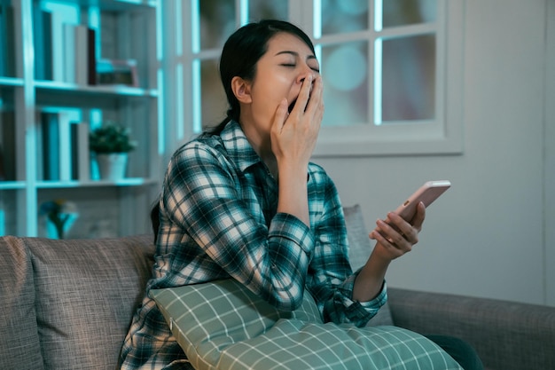 Triste chica asiática con camisa bostezando navegando por sitios web aburridos en un smartphone durante el tiempo libre en un apartamento nocturno. Blogger femenina desanimada leyendo noticias repetitivas pasar tiempo de recreación redes sociales