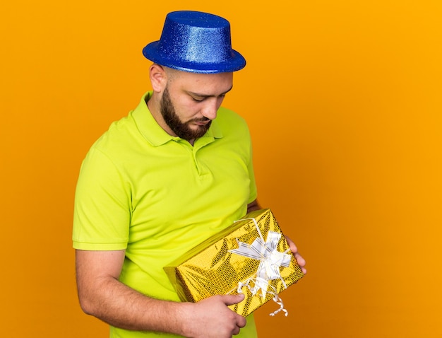 Triste com a cabeça baixa, jovem usando chapéu de festa segurando uma caixa de presente isolada na parede laranja