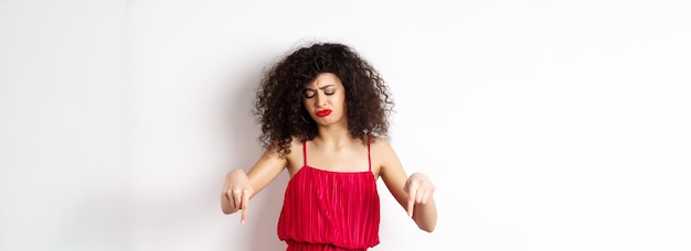 Triste y decepcionada mujer de pelo rizado con vestido rojo frunciendo el ceño y mirando hacia abajo celosa apuntando a pr