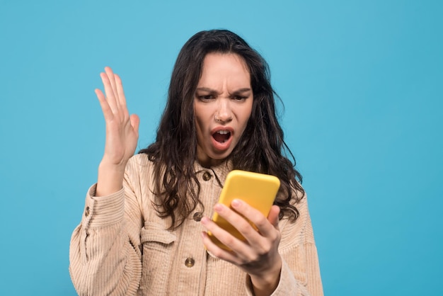 Triste e chocada jovem morena europeia com boca aberta olha para smartphone isolado em azul