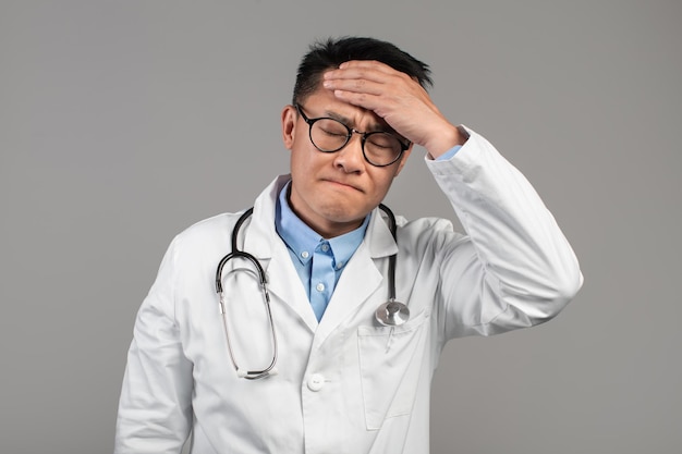 Triste enfermo divertido adulto hombre asiático médico en gafas de bata blanca toca la frente sufre de fiebre