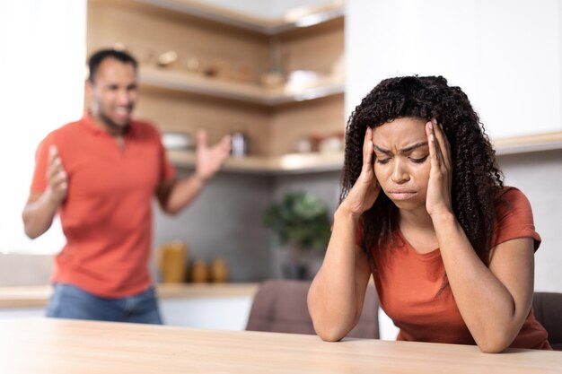 Triste enojado joven marido afroamericano jura a esposa ofendida se sienta a la mesa en el interior de la cocina