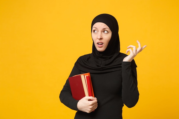 Triste estudante muçulmana árabe em roupas pretas de hijab contém livros isolados no retrato de parede amarela. Estilo de vida religioso de pessoas, educação no conceito de ensino médio. .