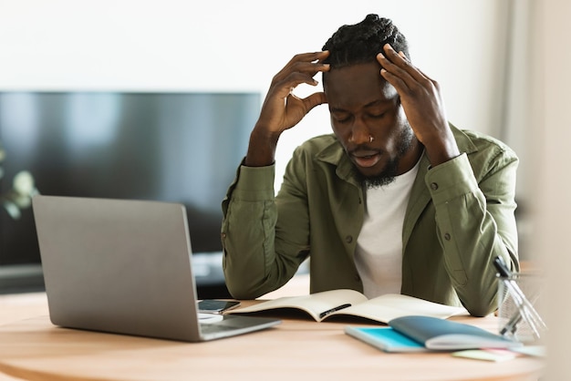 Triste freelancer negro olhando para laptop no local de trabalho sofrendo de dor de cabeça ou tendo problemas