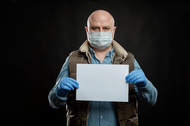 Triste hombre calvo en una máscara médica y guantes con un cartel en blanco en sus manos. Espacio para texto. El desempleo y la crisis mundial durante la pandemia de coronavirus.