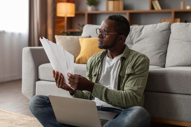 Triste homem negro maduro pensativo em óculos com trabalho de documento de analista de laptop no interior da sala de estar