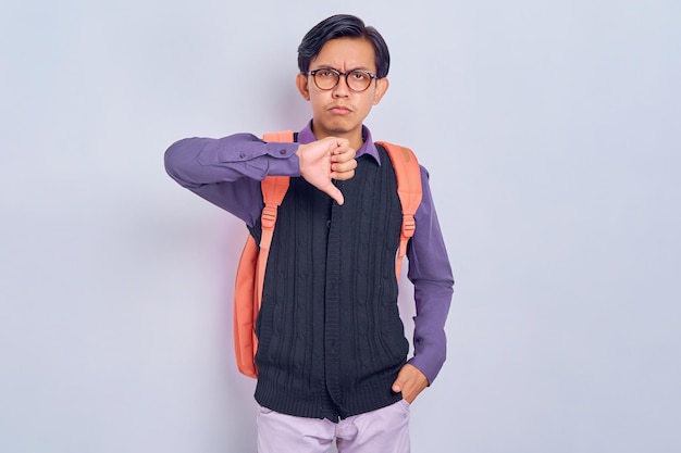 Triste insatisfecho descontento joven estudiante asiático hombre vestido con ropa informal con mochila mostrando el pulgar hacia abajo gesto de disgusto aislado en fondo gris Concepto de universidad de escuela secundaria
