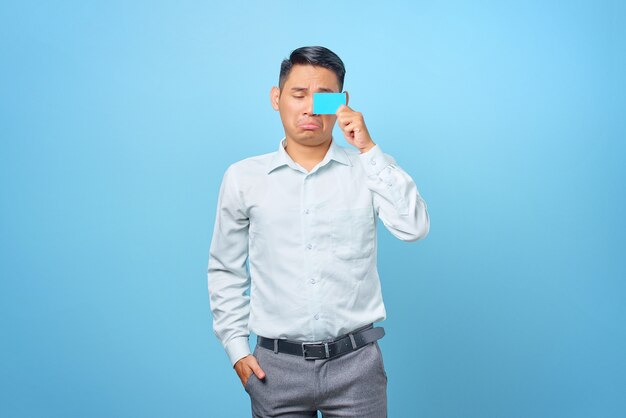 Triste jovem empresário bonito cobrindo os olhos com um cartão de crédito em fundo azul