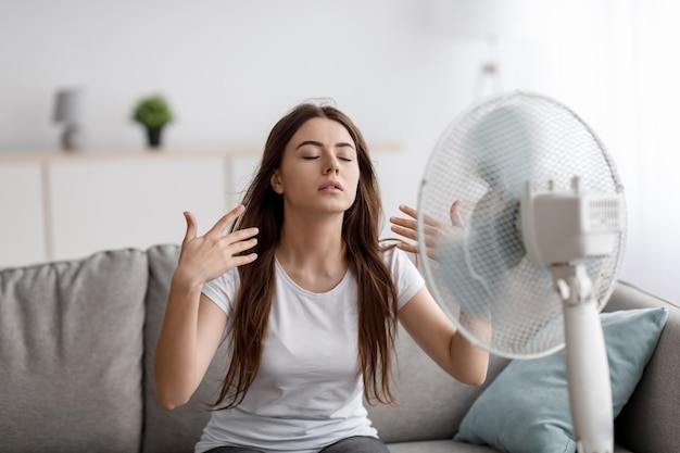 Foto triste jovem europeia que sofre de calor senta-se no sofá pega ar frio do ventilador e acena as mãos