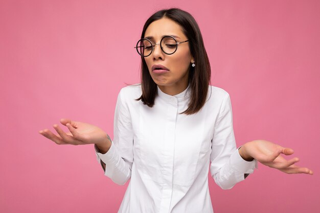 Triste jovem morena atraente vestindo roupas elegantes todos os dias e óculos ópticos modernos, isolados na parede colorida, olhando para baixo e tendo dúvidas.