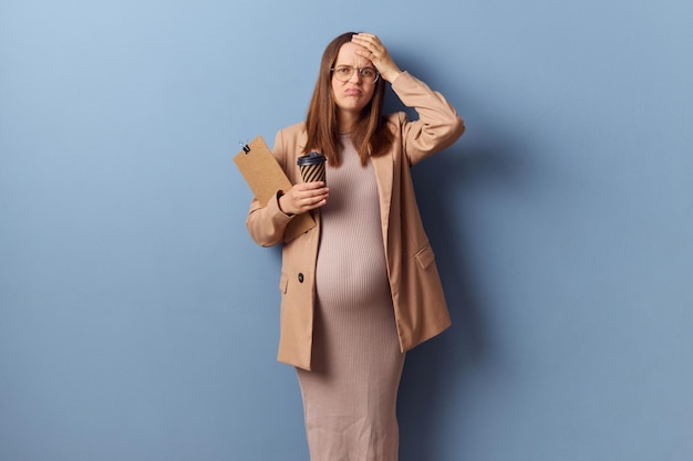 Triste joven adulta infeliz mujer embarazada con vestido y chaqueta posando aislada sobre fondo azul haciendo el gesto de la palma facial teniendo problemas con los documentos tocando su cabeza