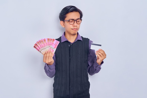 Triste joven asiático con camisa de chaleco que muestra billetes de dinero en rupias y tarjeta de crédito aislado de fondo blanco