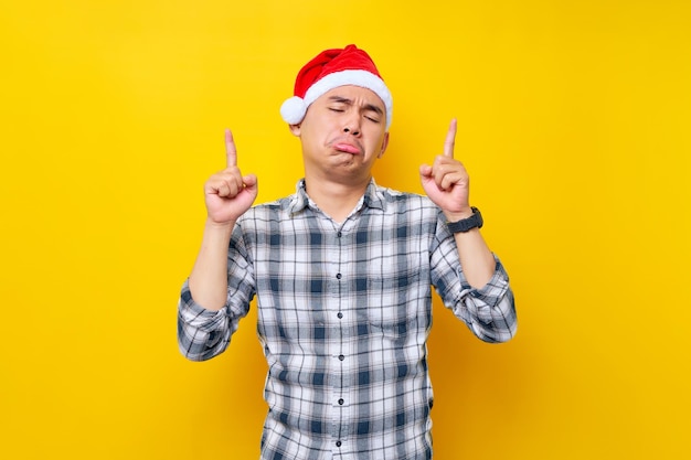 Triste joven asiático con una camisa a cuadros en un sombrero de Navidad señalando con el dedo sobre el área del espacio de trabajo en la celebración de fondo amarillo Navidad y el concepto de Año Nuevo