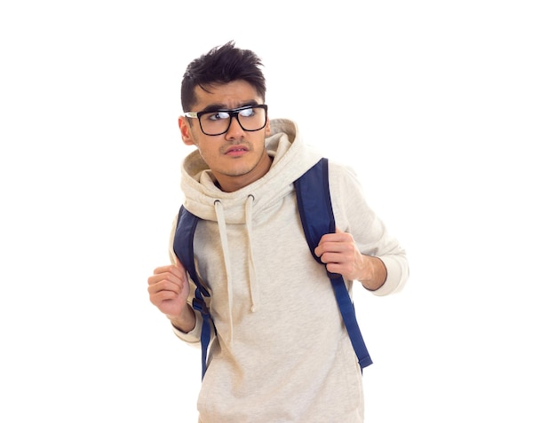 Triste joven con cabello negro en capucha blanca con gafas negras y mochila azul en estudio