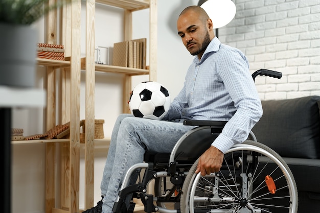 Triste joven discapacitado en silla de ruedas sosteniendo un balón de fútbol