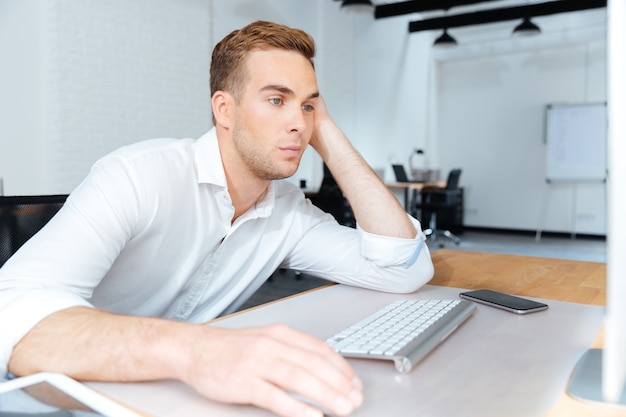 Triste joven empresario aburrido sentado y trabajando con la computadora en la oficina