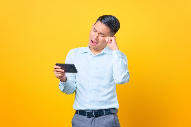 Triste joven empresario guapo jugando un juego en el teléfono móvil sobre fondo amarillo