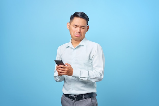 Triste joven empresario guapo sosteniendo y usando smartphone sobre fondo azul.