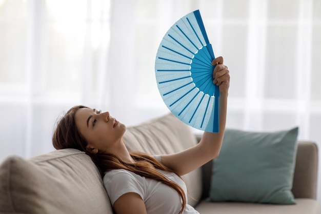 Triste joven europea cansada que sufre de calor en el sofá y se agita el ventilador en la sala de estar