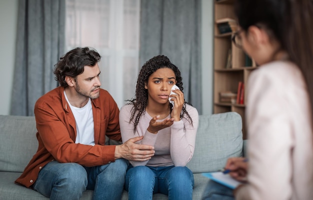 Triste joven europeo macho calma llorando mujer negra hablar con psiquiatra en el interior de la clínica